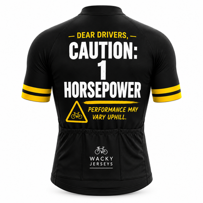 Black short-sleeve cycling jersey shown from the back with bold white and yellow text reading 'DEAR DRIVERS, CAUTION: 1 HORSEPOWER' and a warning triangle graphic. The back pocket area has the Wacky Jerseys logo.