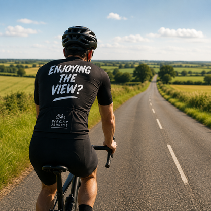 A cyclist viewed from behind riding on a country road, wearing a black jersey with the white text 'ENJOYING THE VIEW?' and 'WAGGY JERSEYS' on the back, with green fields and a blue sky in the background.