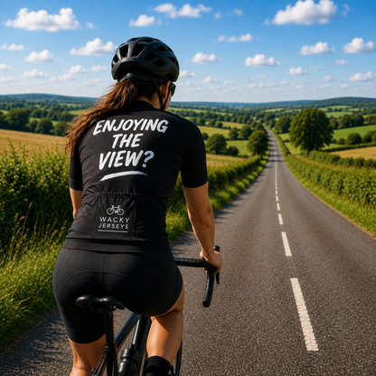 A cyclist viewed from behind riding on a country road, wearing a black cycling jersey with the text 'ENJOYING THE VIEW?' and 'WACKY CYCLES' on the back, along with a black helmet and shorts against a scenic green landscape and blue sky.