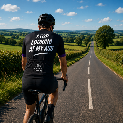 A cyclist viewed from behind riding on a country road, wearing a black cycling jersey with white text reading 'STOP LOOKING AT MY ASS' and a visible 'WACKY JERSEYS' logo on the back pocket, against a bright blue sky and green fields.