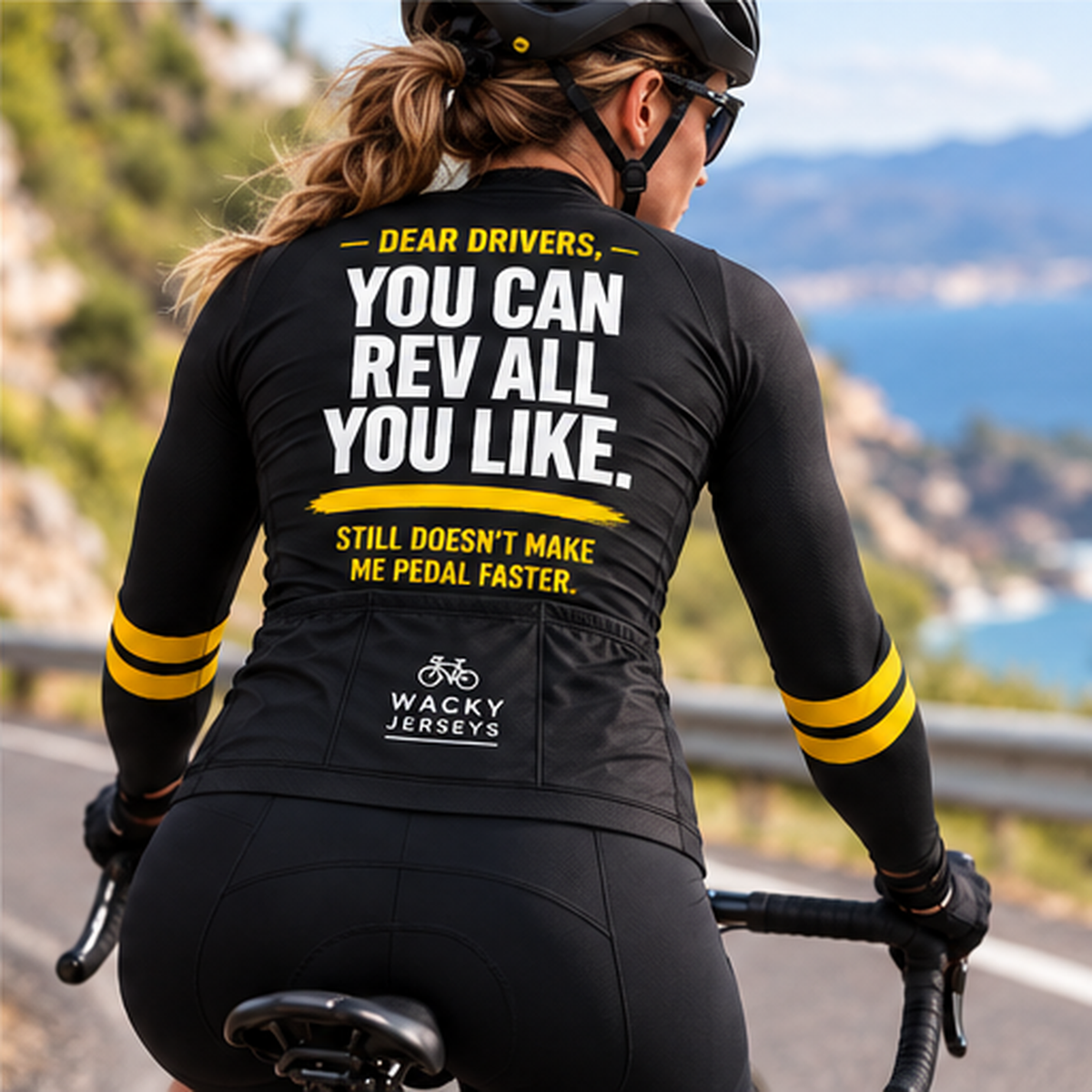 A woman cycling on a road with a scenic coastal landscape in the background, viewed from behind. She is wearing a black long-sleeve cycling jersey with bold yellow and white text on the back and yellow stripes on the sleeves.